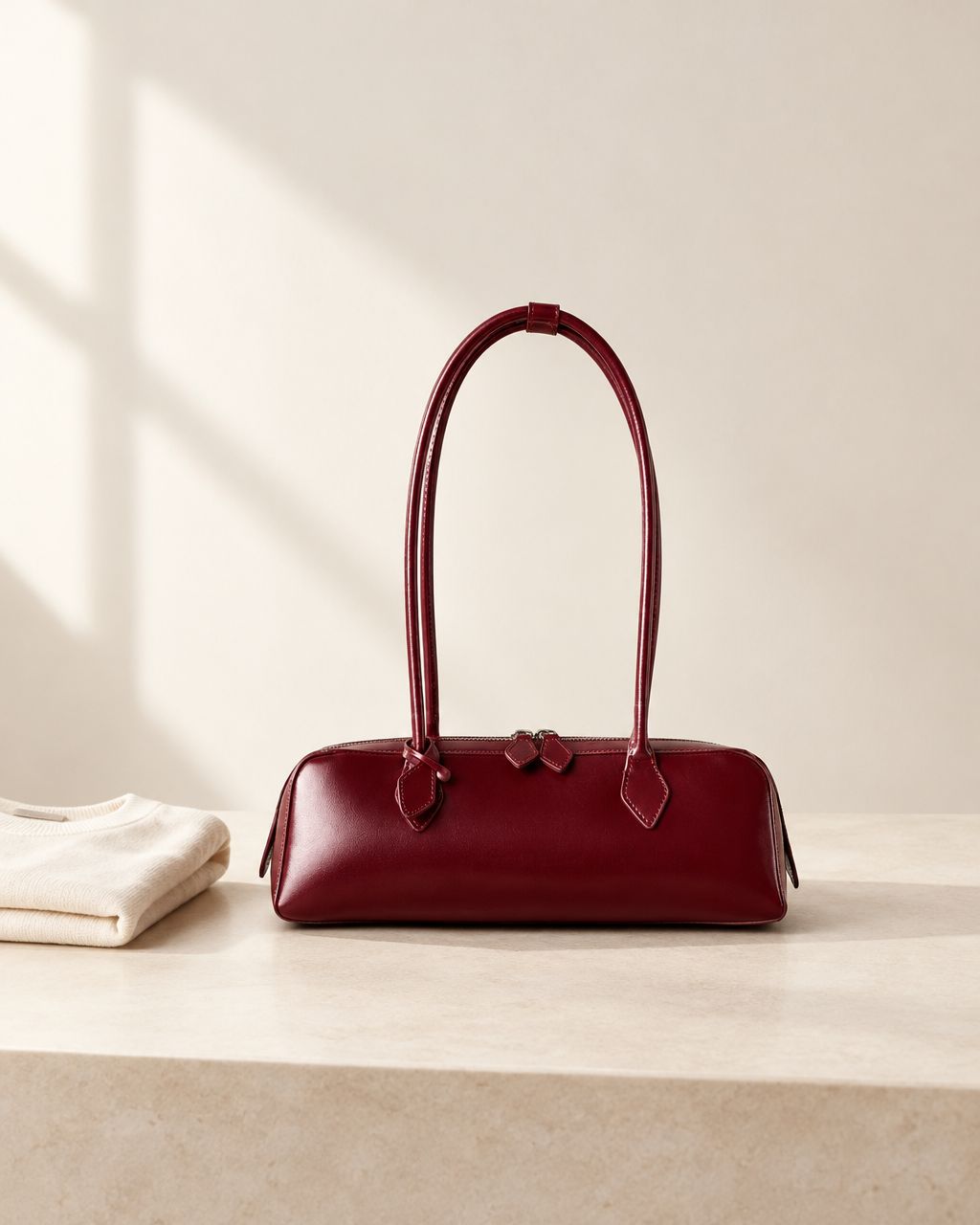 Burgundy leather baguette bag on an ivory tabletop beside a folded cream garment